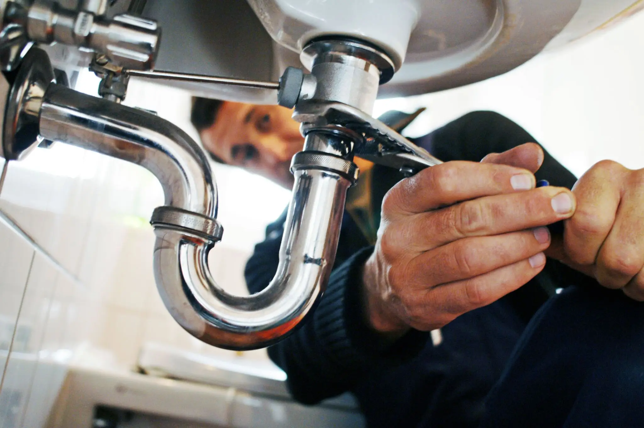 A plumber tightening a pipe under a sink using a wrench.
